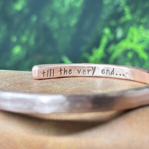 May include: A copper-toned metal cuff bracelet with a hammered texture. The interior surface of the bracelet features the stamped black text "till the very end..." in a simple, lowercase sans-serif font, clearly visible along the inner band.