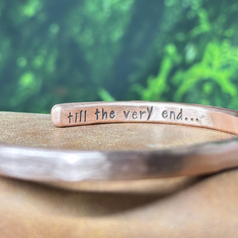 May include: A copper-toned metal cuff bracelet with a hammered texture. The interior surface of the bracelet features the stamped black text "till the very end..." in a simple, lowercase sans-serif font, clearly visible along the inner band.