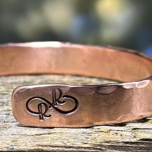 May include: A copper cuff bracelet with a hammered texture. The bracelet features an engraved design of the letters "R" and "B" in a cursive font. The bracelet is displayed on a wooden surface.