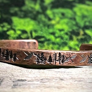 May include: Copper cuff bracelet with a mountain and forest design. The bracelet features a detailed etched landscape of trees and mountains. The cuff is a warm brown color and has a rustic, handcrafted appearance.