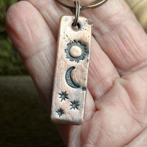 May include: A copper-colored rectangular pendant with a sun, crescent moon, and three stars design. The pendant is attached to a copper-colored ring. The sun has a detailed, textured design, and the moon and stars are also detailed.