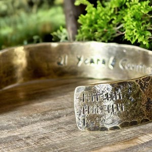 May include: A hammered brass cuff bracelet with the inscription "21 Years Counting" and tally marks. The open-ended bracelet is displayed on a wooden surface with a blurred green background.