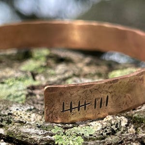 May include: A copper cuff bracelet with a hammered texture. The bracelet has an open design and features etched tally marks. The warm, metallic tones of the bracelet contrast with the natural background, highlighting the handcrafted details.