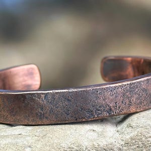 Puede incluir: Un brazalete de cobre con superficie texturizada. El brazalete abierto tiene forma rectangular y un aspecto oscuro y envejecido. El metal tiene un tono marrón rojizo.