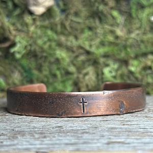 Puede incluir: Una pulsera de puño de cobre con un diseño de cruz estampado. La pulsera tiene un aspecto rústico y desgastado y está sobre un fondo borroso verde y marrón. La pulsera mide aproximadamente 1,9 cm de ancho.