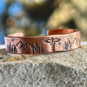 Puede incluir: Brazalete de puño de cobre con un diseño de montaña y árboles. El brazalete también presenta un símbolo médico. El brazalete es abierto y tiene un acabado rústico y texturizado.