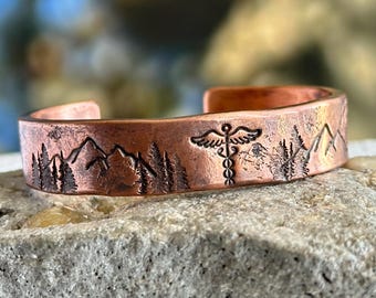 Puede incluir: Brazalete de puño de cobre con un diseño de montaña y árboles. El brazalete también presenta un símbolo médico. El brazalete es abierto y tiene un acabado rústico y texturizado.