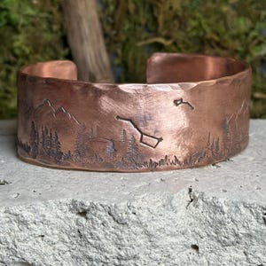 May include: A copper cuff bracelet with an etched landscape design featuring mountains, trees, and constellations. The bracelet has a hammered texture and a rustic aesthetic. The design includes the Big Dipper and other stars.