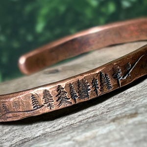 Puede incluir: Brazalete de cobre con un diseño detallado de bosque y montaña. El brazalete presenta un paisaje grabado de árboles y una cordillera. El metal tiene un acabado rústico y texturizado, evocando un tema natural y al aire libre.