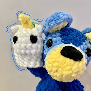 May include: A blue crocheted stuffed animal holding a white crocheted unicorn with a yellow horn. The stuffed animal has a yellow nose and black eyes.