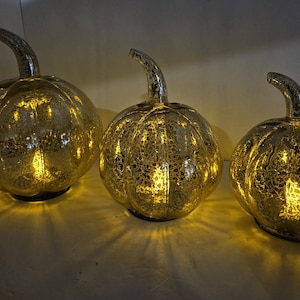 May include: Three decorative, illuminated pumpkin-shaped glass lanterns. Each pumpkin has a silver stem and a textured, crackled surface. The pumpkins are lit from within, casting a warm, golden glow, perfect for fall or Halloween decor.