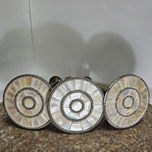 May include: Three decorative candle holders with circular, mother-of-pearl designs. Each holder features concentric circles of iridescent shell segments, framed by a silver-toned metal. The holders are arranged on a textured, brown surface.