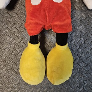 Op de afbeelding: Een pluche Mickey Mouse pop met een rode broek, witte knopen, zwarte benen en grote gele schoenen. De pop heeft witte handschoenen en zwarte oren. Het speelgoed is ontworpen voor kinderen.