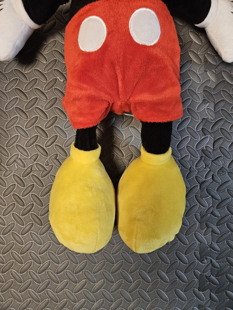 Op de afbeelding: Een pluche Mickey Mouse pop met een rode broek, witte knopen, zwarte benen en grote gele schoenen. De pop heeft witte handschoenen en zwarte oren. Het speelgoed is ontworpen voor kinderen.