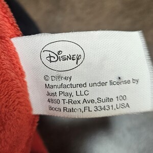 Op de afbeelding: Een wit label met het Disney-logo en tekst. Het label luidt "&copy; Disney, Vervaardigd onder licentie door Just Play, LLC, 4850 T-Rex Ave, Suite 100, Boca Raton, FL 33431, USA." Het label bevindt zich naast een rood pluche materiaal.
