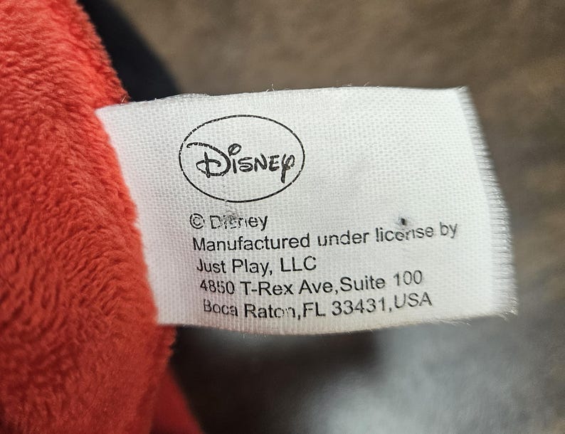 Op de afbeelding: Een wit label met het Disney-logo en tekst. Het label luidt "&copy; Disney, Vervaardigd onder licentie door Just Play, LLC, 4850 T-Rex Ave, Suite 100, Boca Raton, FL 33431, USA." Het label bevindt zich naast een rood pluche materiaal.