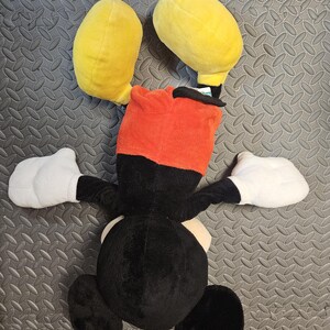 Op de afbeelding: Een pluche speelgoed van een stripfiguur, met grote gele oren, een rode en zwarte kop, witte handschoenen en een zwart lichaam. Het speelgoed ligt op een getextureerd grijs oppervlak.