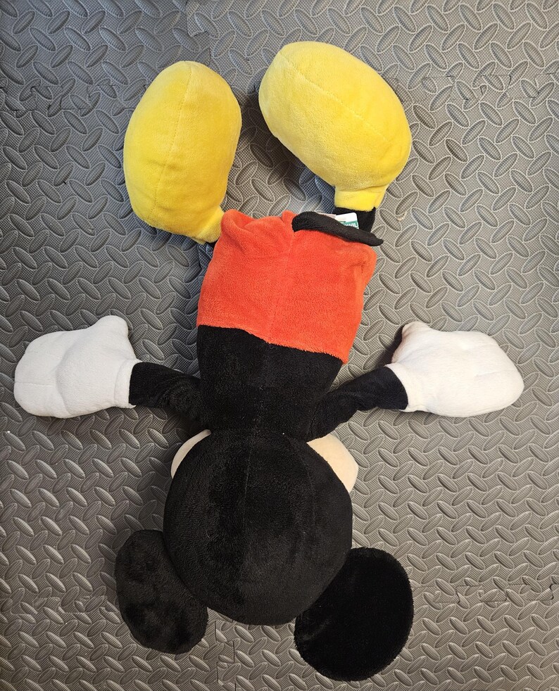 Op de afbeelding: Een pluche speelgoed van een stripfiguur, met grote gele oren, een rode en zwarte kop, witte handschoenen en een zwart lichaam. Het speelgoed ligt op een getextureerd grijs oppervlak.