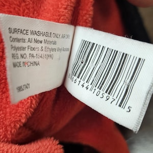 Op de afbeelding: Close-up van een wit label met zwarte tekst en een barcode, bevestigd aan een rood pluche materiaal. Het label luidt "SURFACE WASHABLE ONLY. AIR DRY" en "Contents: All New Materials".