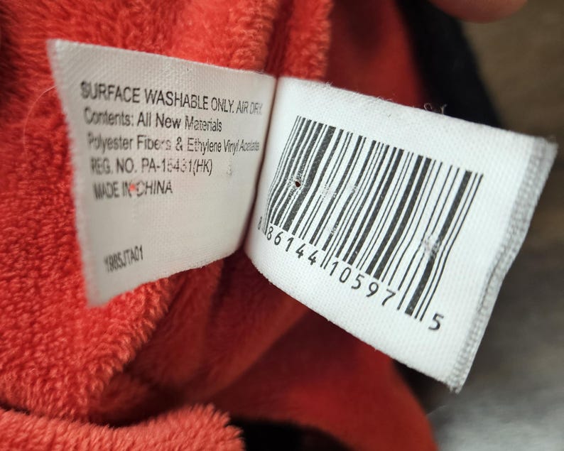 Op de afbeelding: Close-up van een wit label met zwarte tekst en een barcode, bevestigd aan een rood pluche materiaal. Het label luidt "SURFACE WASHABLE ONLY. AIR DRY" en "Contents: All New Materials".