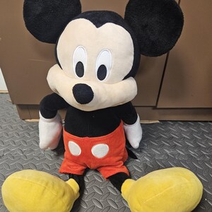 Op de afbeelding: Een Mickey Mouse knuffel. Het speelgoed heeft grote zwarte oren, een zwarte body, een rode korte broek met witte knopen, witte handschoenen en grote gele schoenen. Het gezicht is wit met zwarte ogen en een zwarte neus.