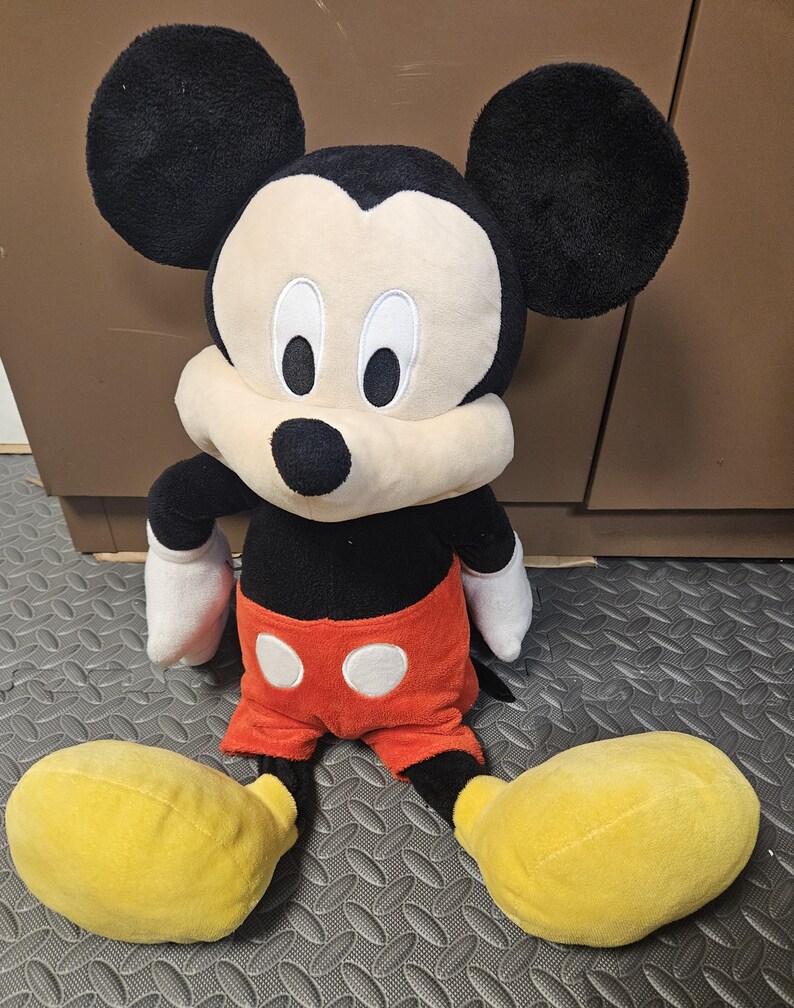 Op de afbeelding: Een Mickey Mouse knuffel. Het speelgoed heeft grote zwarte oren, een zwarte body, een rode korte broek met witte knopen, witte handschoenen en grote gele schoenen. Het gezicht is wit met zwarte ogen en een zwarte neus.