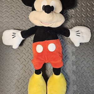 Op de afbeelding: Een pluche Mickey Mouse pop met een zwart lichaam, oren en armen, een cr&egrave;mekleurig gezicht en witte handschoenen. De pop heeft een oranje broek met witte knopen en grote gele schoenen. De pop is ongeveer 61 cm hoog.