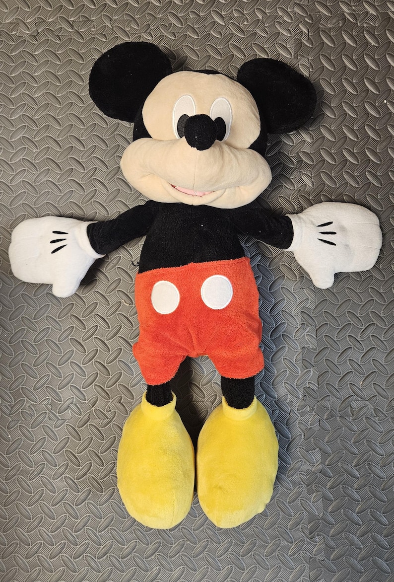 Op de afbeelding: Een pluche Mickey Mouse pop met een zwart lichaam, oren en armen, een cr&egrave;mekleurig gezicht en witte handschoenen. De pop heeft een oranje broek met witte knopen en grote gele schoenen. De pop is ongeveer 61 cm hoog.