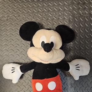 Op de afbeelding: Een pluche Mickey Mouse-speelgoed met een zwart lichaam, grote zwarte oren en een cr&egrave;mekleurig gezicht. Het speelgoed heeft witte handschoenen met drie zwarte lijnen op elke hand, een rode broek met twee witte knopen en staat in een uitnodigende pose.