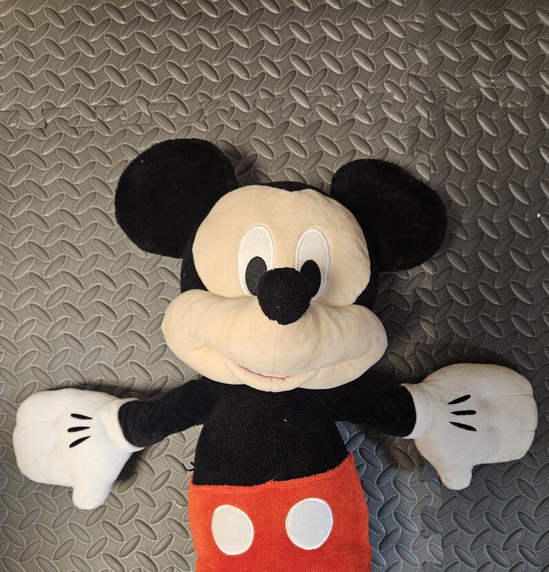 Op de afbeelding: Een pluche Mickey Mouse-speelgoed met een zwart lichaam, grote zwarte oren en een cr&egrave;mekleurig gezicht. Het speelgoed heeft witte handschoenen met drie zwarte lijnen op elke hand, een rode broek met twee witte knopen en staat in een uitnodigende pose.