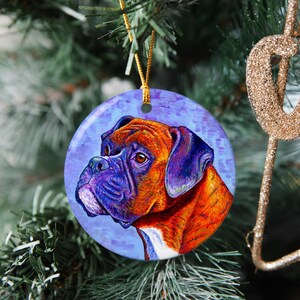 May include: A round ceramic Christmas ornament featuring a colorful painting of a Boxer dog with a blue, purple, and orange color scheme. The dog is looking to the left of the image.