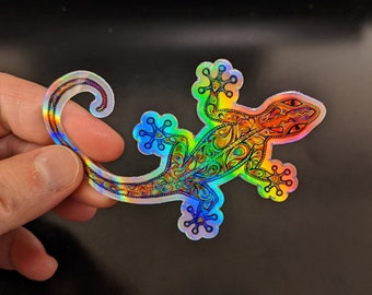 May include: A holographic sticker of a gecko with a colorful, intricate design. The gecko is in a curled position with its tail wrapped around its body.