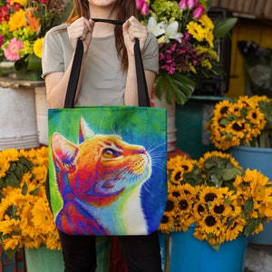 May include: A colorful tote bag with a vibrant orange cat illustration. The cat is looking up with its mouth closed. The background is a mix of green, blue, and purple.