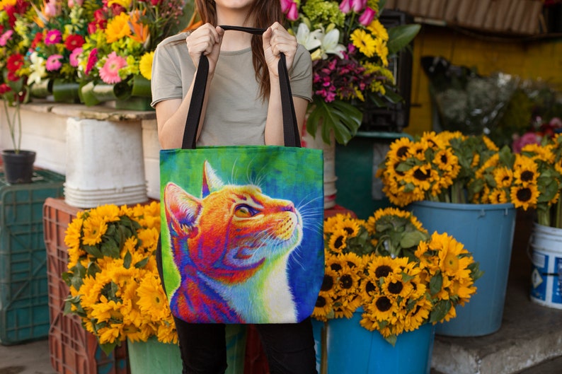May include: A colorful tote bag with a vibrant orange cat illustration. The cat is looking up with its mouth closed. The background is a mix of green, blue, and purple.