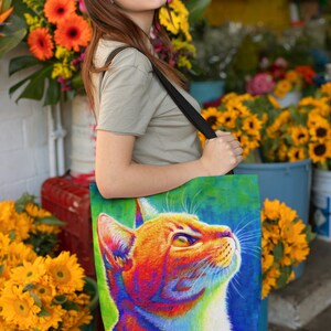 May include: A woman is carrying a tote bag with a colorful illustration of a cat's head. The cat has orange, red, yellow, blue, and green fur. The bag has a black strap.