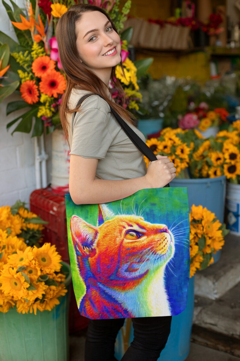 May include: A woman is carrying a tote bag with a colorful illustration of a cat's head. The cat has orange, red, yellow, blue, and green fur. The bag has a black strap.
