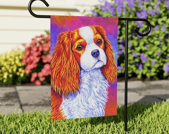 May include: A colorful garden flag featuring a close-up portrait of a Cavalier King Charles Spaniel dog with a white and red coat. The dog's fur is painted in a vibrant, almost psychedelic style with shades of orange, red, pink, and purple.