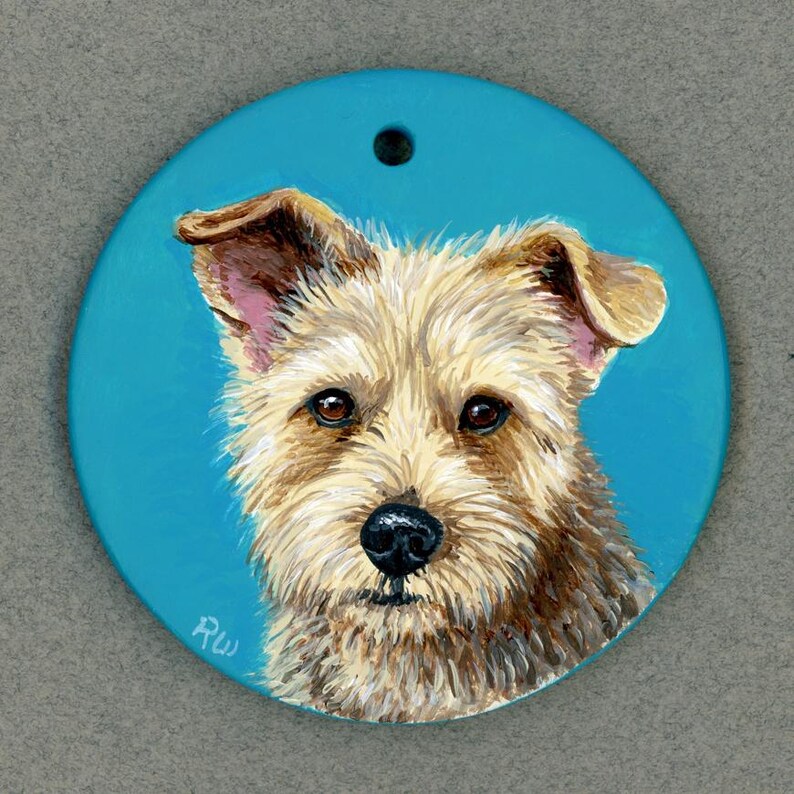May include: A hand-painted ceramic ornament featuring a portrait of a small, light brown dog with a fluffy coat. The dog has brown eyes and a black nose. The ornament is painted on a bright blue background and has a hole at the top for hanging.