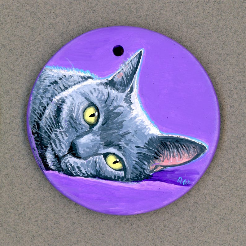 May include: A hand-painted ceramic ornament featuring a gray cat with yellow eyes, lying on a purple background. The cat's head is tilted to the side, and its eyes are looking directly at the viewer. The ornament is round and has a hole at the top for hanging.