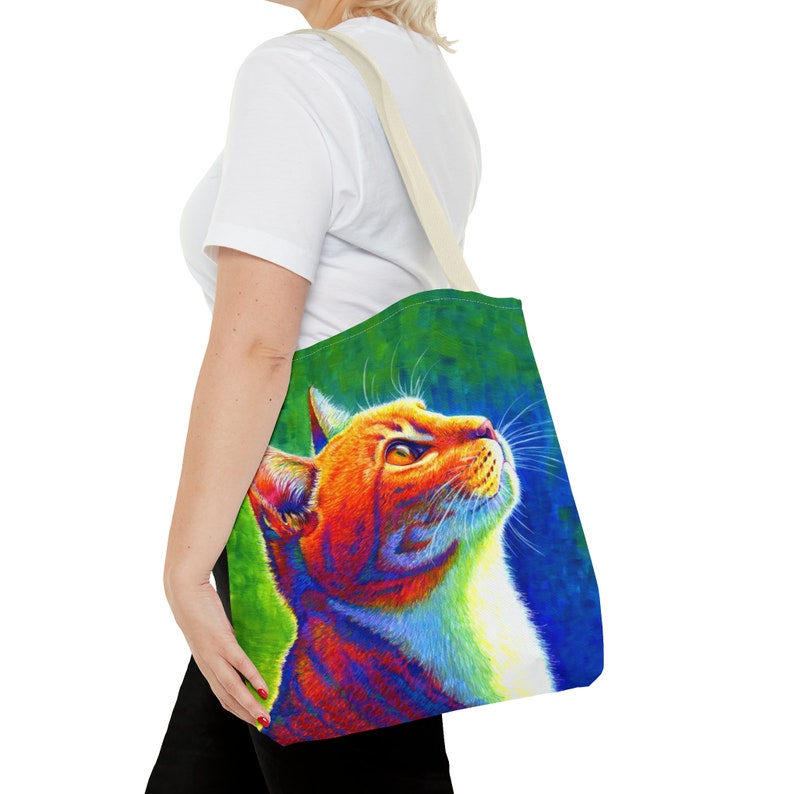 May include: A tote bag with a colorful illustration of a cat looking up. The cat is orange, white, and has a rainbow-like effect. The background is a green and blue gradient.