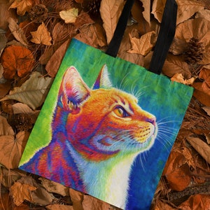 May include: A tote bag with a colorful illustration of a cat's face. The cat has orange, red, yellow, blue, and white fur. The background is a gradient of green, blue, and purple.