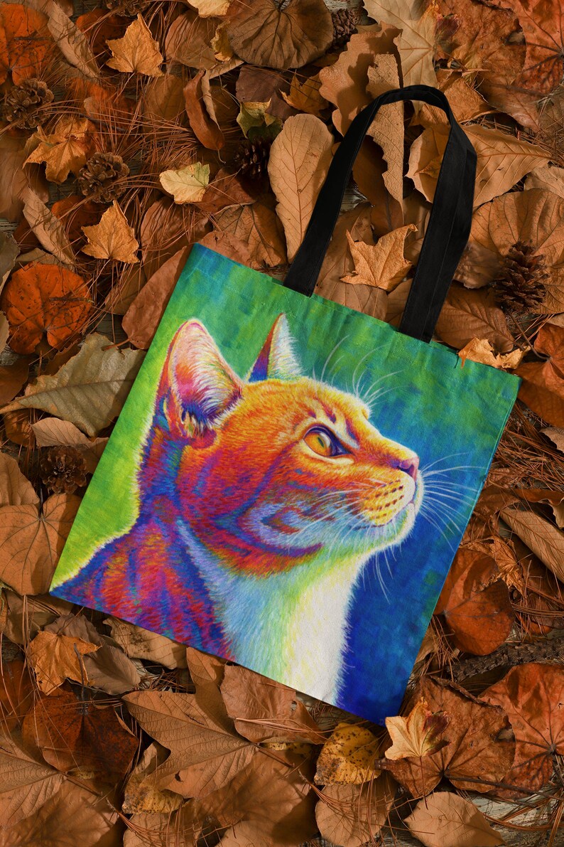 May include: A tote bag with a colorful illustration of a cat's face. The cat has orange, red, yellow, blue, and white fur. The background is a gradient of green, blue, and purple.