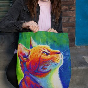 May include: A tote bag with a colorful illustration of a cat's head. The cat has orange fur with a white chest and is looking up. The background is a vibrant green and blue.
