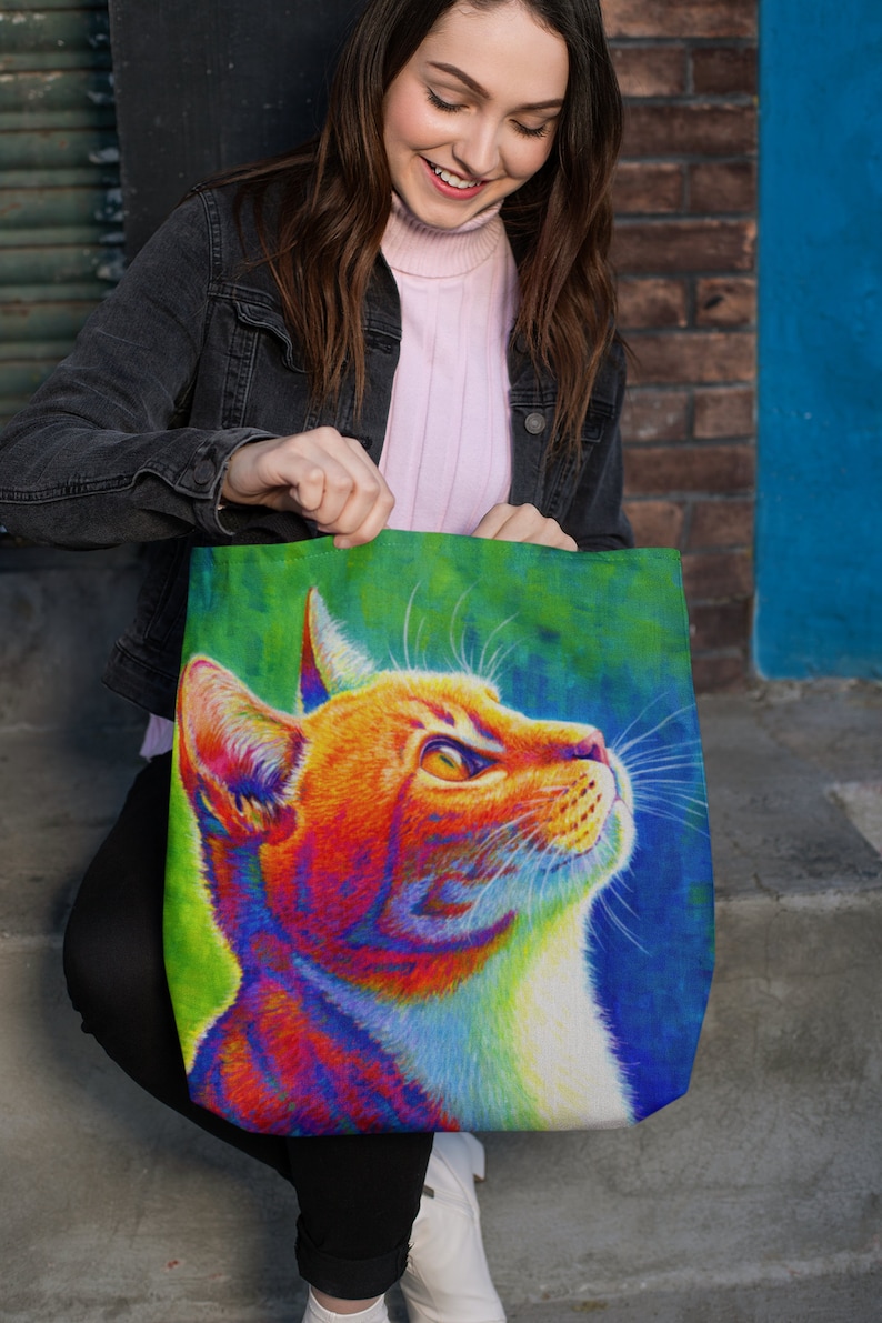 May include: A tote bag with a colorful illustration of a cat's head. The cat has orange fur with a white chest and is looking up. The background is a vibrant green and blue.