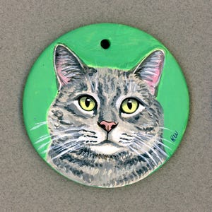 May include: A hand-painted ceramic ornament featuring a gray tabby cat with green eyes. The cat is painted on a green background and the ornament has a hole at the top for hanging.