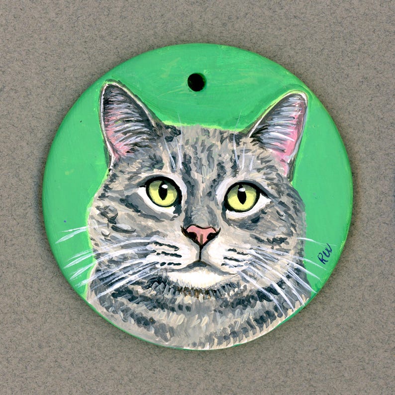 May include: A hand-painted ceramic ornament featuring a gray tabby cat with green eyes. The cat is painted on a green background and the ornament has a hole at the top for hanging.