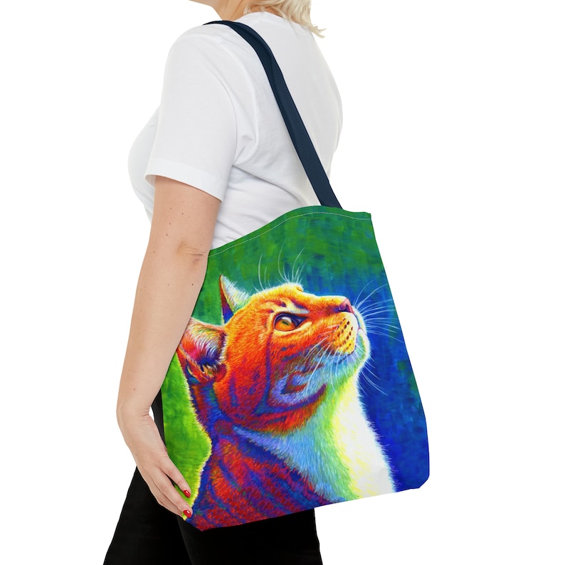 May include: A tote bag with a colorful illustration of a cat looking up. The cat is orange, white, and has a rainbow effect. The background is green and blue.