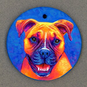 May include: A hand-painted ceramic ornament featuring a close-up portrait of a boxer dog with a bright orange, yellow, and white fur pattern. The dog has a blue nose and a bright blue background. The ornament is round and has a hole at the top for hanging.