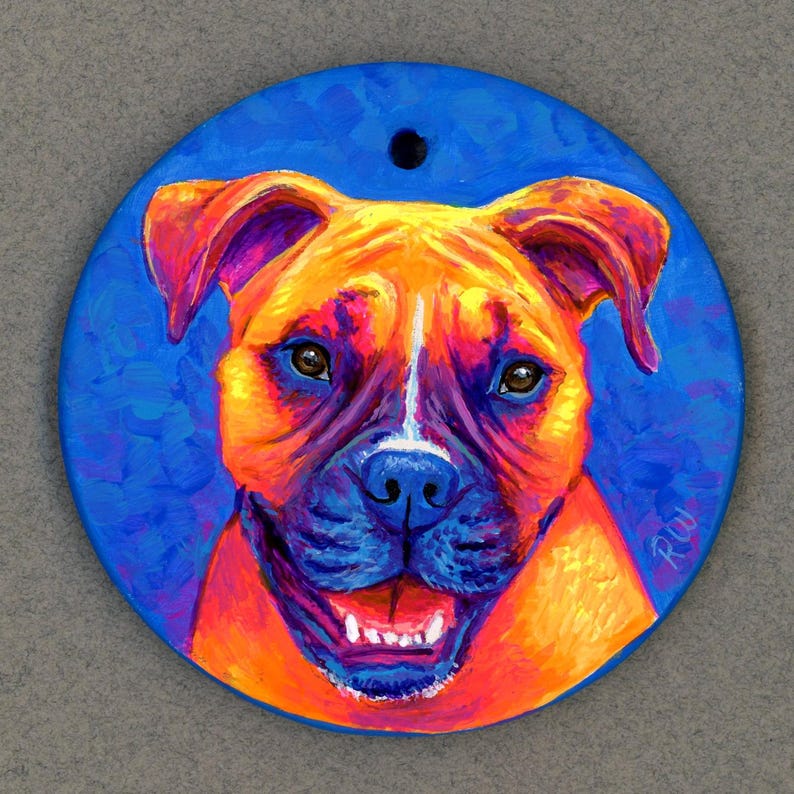May include: A hand-painted ceramic ornament featuring a close-up portrait of a boxer dog with a bright orange, yellow, and white fur pattern. The dog has a blue nose and a bright blue background. The ornament is round and has a hole at the top for hanging.