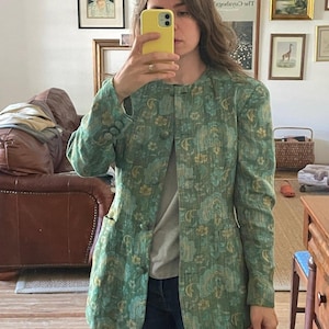 May include: A long-sleeved, collarless blazer in a textured green fabric featuring a repeating floral and abstract pattern in shades of yellow and light green. The jacket has a button-front closure with matching fabric-covered buttons and two front pockets.